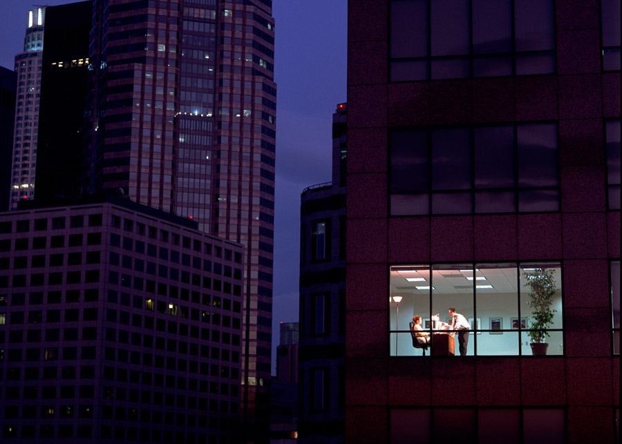 long shot of a city skyline at night, with one business office lit up to show a man and woman in a conference room
